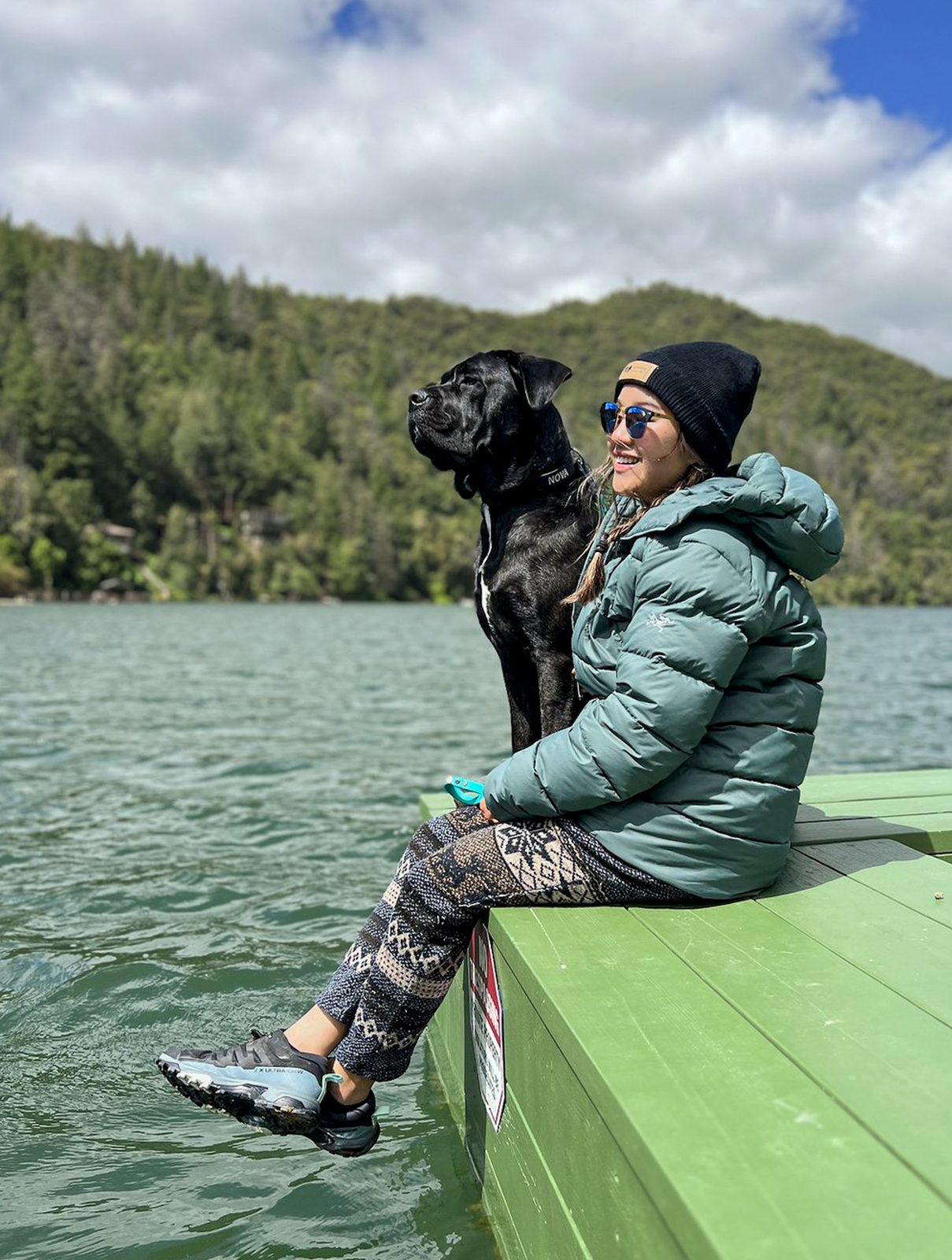 Jasmine smiling on a green dock by a mountain lake with Nova, their Cane Corso, beside her